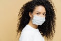 Photo of dark skinned Afro American woman with curly bushy hair, wears protective mask during coronavirus outbreak, isolated on