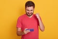 Photo of delighted man dresses red casual t shirt playing video game on cellphone and clenching fist isolated over yellow studio