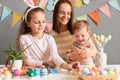 Photo of delighted positive female with brown hair and her children painting eggs, preparing for Easter, spending time together,