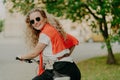 Photo of delighted woman poses back at camera, rides bicycle outdoor, wears sunglasses, t shirt and sweater on shoulders, poses