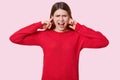 Photo of desperate Caucasian woman pluggs ears with fingers, shouts angrily, dressed in red jumper, irritated with annoying sound