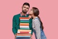 Photo of displeased unshaven man carries textbooks, recieves kiss, wears spectacles, isolated over pink background