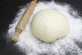 A photo of dough, rolling pin and wheat white flour sprinkled on black wooden table. Selective soft focus. Dough ready for cooking