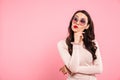 Photo of dreaming adult girl with red lips wearing trendy sunglasses looking aside touching chin with hand, isolated over pink ba Royalty Free Stock Photo