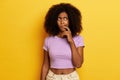 Photo of dreamy dark skinned young woman with afro hair, looks thoughtfully aside, keeps finger near underlip, dressed Royalty Free Stock Photo