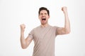 Photo of ecstatic guy 30s in beige t-shirt shouting as celebrating his triumph with clenched fists, isolated over white background Royalty Free Stock Photo