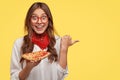 Photo of emotive surprised brunette lady with toothy smile, wears red bandana, holds slice of pizza, points with thumb