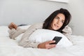 Photo of european woman 30s using mobile phone, while lying in bed with white linen in bright room Royalty Free Stock Photo
