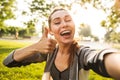 Photo of feminine fitness girl 20s in sportswear wearing bluetooth earpod taking selfie and showing thumb up, while walking in gr Royalty Free Stock Photo