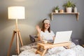 Photo of friendly happy smiling Caucasian woman wearing sleep mask sitting with laptop in bed at home and having video call, Royalty Free Stock Photo