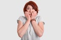 Photo of frightened red haired woman bites fingers nails, looks with nervous expression, afraids of difficulties, dressed in