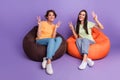 Photo of funky cheerful girls dressed t-shirts sitting bean bags showing v-signs isolated purple color background Royalty Free Stock Photo