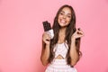 Photo of funny woman 20s wearing dress smiling and eating chocolate bar, isolated over pink background Royalty Free Stock Photo