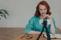 Photo of ginger female blogger shoots video of herself for personal blog, looks at camera of smartphone on tripod, chats with Royalty Free Stock Photo