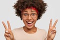 Photo of glad Afro American lady shows victory or peace sign, gestures with both hands, smiles broadly, wears round