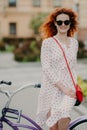 Photo of glad red haired woman with gentle smile, spends leisure time riding bicycle on city streets during sunny windy day, wears