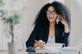 Photo of glad smiling dark skinned woman calls business partner, being in good mood, makes notes, sits at workplace, wears optical