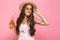 Photo of glamour woman 20s wearing sunglasses and straw hat drinking lemonade from glass bottle, isolated over pink background Royalty Free Stock Photo