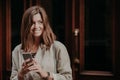 Photo of good looking woman searches information, uses cell phone, dressed in elegant jacket, poses against door background, looks