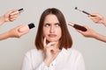 Photo of hands of several beauticians holding their respective equipment giving makeup to attractive woman, standing with pensive