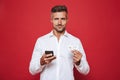 Photo of handsome man in white shirt holding credit card and smartphone, isolated over red background Royalty Free Stock Photo