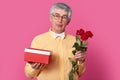 Photo of handsome of mature man in spectacles, dressed in yellow shirt with bowtie, carries red box with present and roses, wants