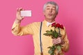 Photo of handsome senior man holds mobilephone in front, makes selfie portrait, pouts lips at camera, wears elegant shirt with