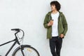 Photo of handsome youngster with curly bushy hair, dressed in white t shirt, trousers, green anorak, keeps hand in pocket, concent