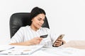 Photo of happy brunette businesswoman 30s holding smartphone and plastic credit card while sitting in armchair at table in office Royalty Free Stock Photo