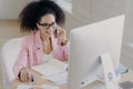 Photo of happy curly haired female wears optical glasses, pink jacket, looks attentively at computer display, sits at desktop with