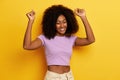 Photo of happy dancing dark skinned attractive woman with hands raised, celebrates her success with satisfied smile