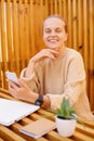 Photo of happy winsome smiling happy Caucasian woman with bun hairstyle wearing beige sweater working on laptop and holding smart Royalty Free Stock Photo