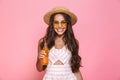 Photo of happy woman 20s wearing sunglasses and straw hat drinking juice from glass bottle, isolated over pink background Royalty Free Stock Photo