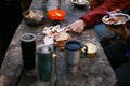 Photo of hiker`s breakfast wooden table with bread, bacon , cans, other meals and hot mugs at the forest camp. People