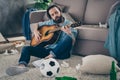 Photo of hipster guy with long beard sitting carpet near sofa holding guitar don`t mind chaos after stag party messy