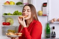 Photo of hungry young cute woman eats with appetite tasty sausage sandwhich, comes after work, stands near opened refrigerator, cl Royalty Free Stock Photo