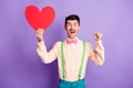 Photo of impressed lucky young man dressed yellow shirt holding large red paper heart rising fist isolated purple color Royalty Free Stock Photo