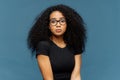 Photo of indignant puzzled Afro American woman looks surprisingly at camera, wears casual black t shirt and spectacles, poses