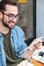 Photo of intelligent young man smiling and reading newspaper while sitting in city cafe outdoors