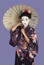 Portrait of Japanese woman in kimono with painted face holding parasol against purple background