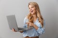 Photo of joyful blonde woman 20s dressed in shirt smiling while holding and pointing finger on silver laptop