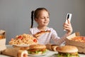 Photo of joyful little girl with pigtails sitting at table isolated over gray background, using mobile phone, having video call,