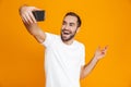 Photo of joyous guy 30s in casual wear laughing and taking selfie on cell phone, isolated over yellow background