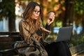 Photo of joyous woman working on laptop while sitting on bench in sunlit alley