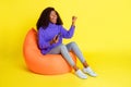 Photo of lady sit beanbag hold gamepad raise fist open mouth wear purple sweater jeans shoes isolated yellow color