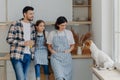 Photo of little female child, her mother and father stand together at modern kitchen, look with funny expressions at jack russell