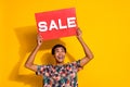 Photo of looking above head young promoter man in trendy t shirt holding red poster sale word isolated on yellow color