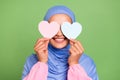 Photo of lovely muslimah woman cover eyes small heart shape love feelings isolated over green color background Royalty Free Stock Photo