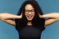 Photo of lovely smiling Afro female with crsip hair, covers ears, being in good mood, wears casual black t shirt, amused by loud