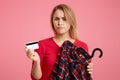 Photo of lovely woman with discontent expression goes shopping in fashionable boutique, chooses outfit, holds plastic card, has no